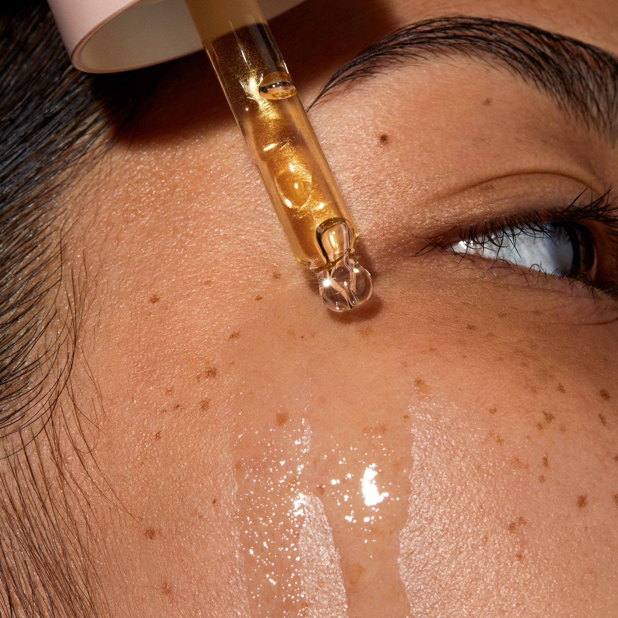 A super close up of model's face Queen oil is applied to her skin with a pipette. The skin is glowy and hydrated.